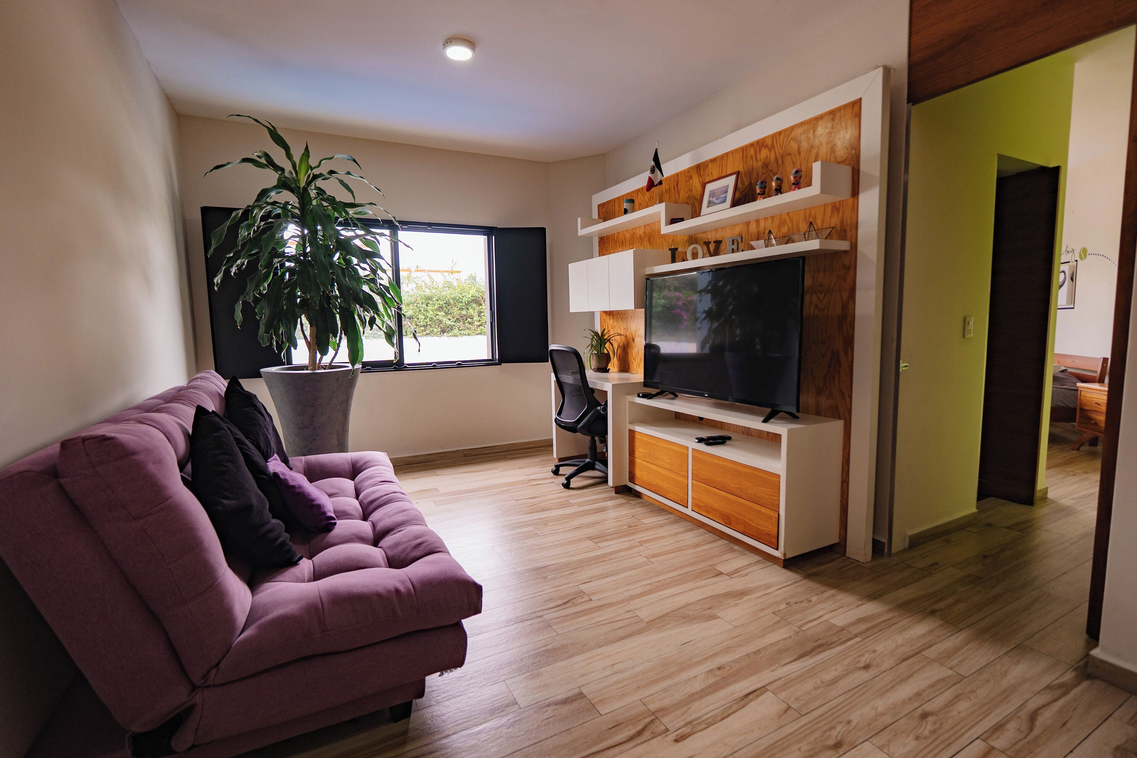 Un salón con un sofá morado, un televisor grande, una planta en maceta y suelo de madera.