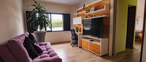 Un salón con un sofá morado, un televisor grande, una planta en maceta y suelo de madera.