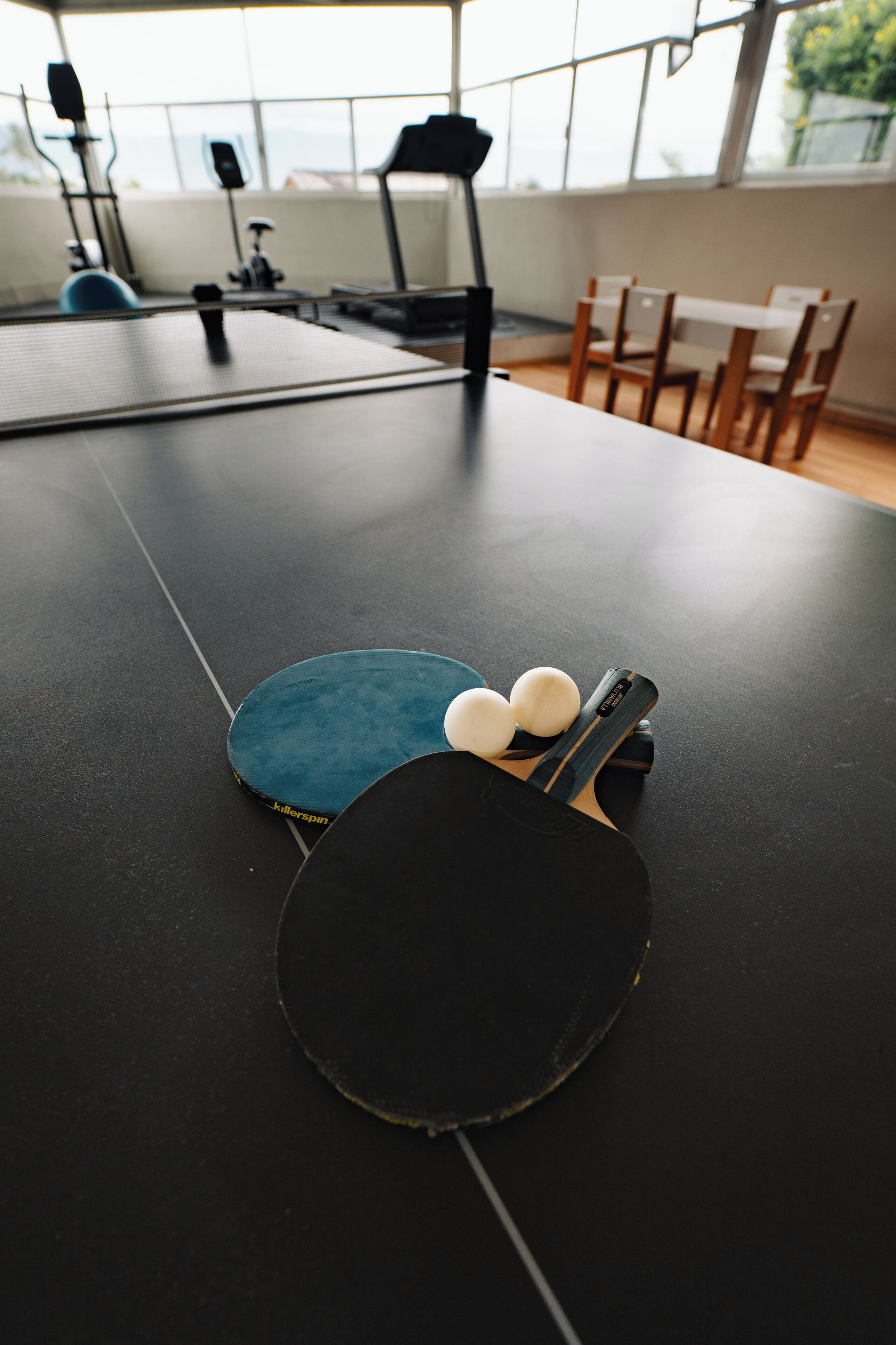 Ett bordtennisbord med två racketar och två bollar i ett rum med träningsutrustning och utsikt genom stora fönster.