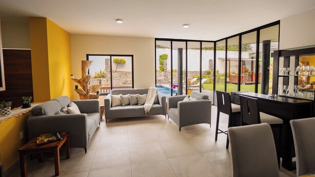A modern living room with a grey sofa set, a coffee table, and a dining area with chairs and a glass-top table. There is a large window with a view of a swimming pool and greenery outside.