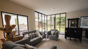 A modern living room with grey sofas, a coffee table, and a TV stand. Large windows offer a view of a garden and outdoor seating area.