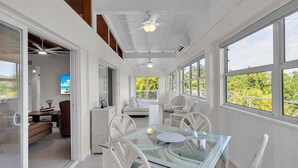 A bright, open-plan living area with a glass dining table surrounded by white wicker chairs, a white sofa, and a ceiling fan. Large windows provide views of greenery outside. - Marathon Beach House | Pool, Dock & Ocean Views (Marathon)