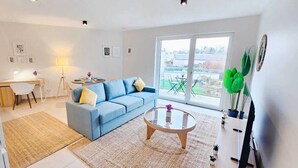 A modern living room with a blue sofa, a glass coffee table, and a large window offering a view of the outdoors. - Warm&Bright 1BR w terrace&parking (Luxembourg City)
