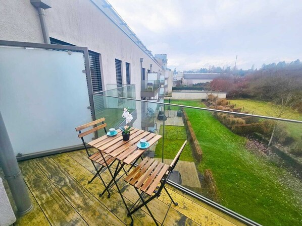 Un balcon moderne avec une table et des chaises en bois, une balustrade en verre et une vue sur un paysage verdoyant.