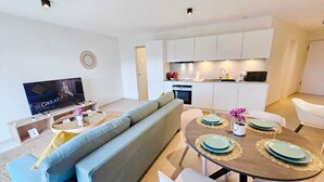 A modern living room with a sofa, a TV, a dining table set with plates and glasses, and a kitchen in the background. - Warm&Bright 1BR w terrace&parking (Luxembourg City)
