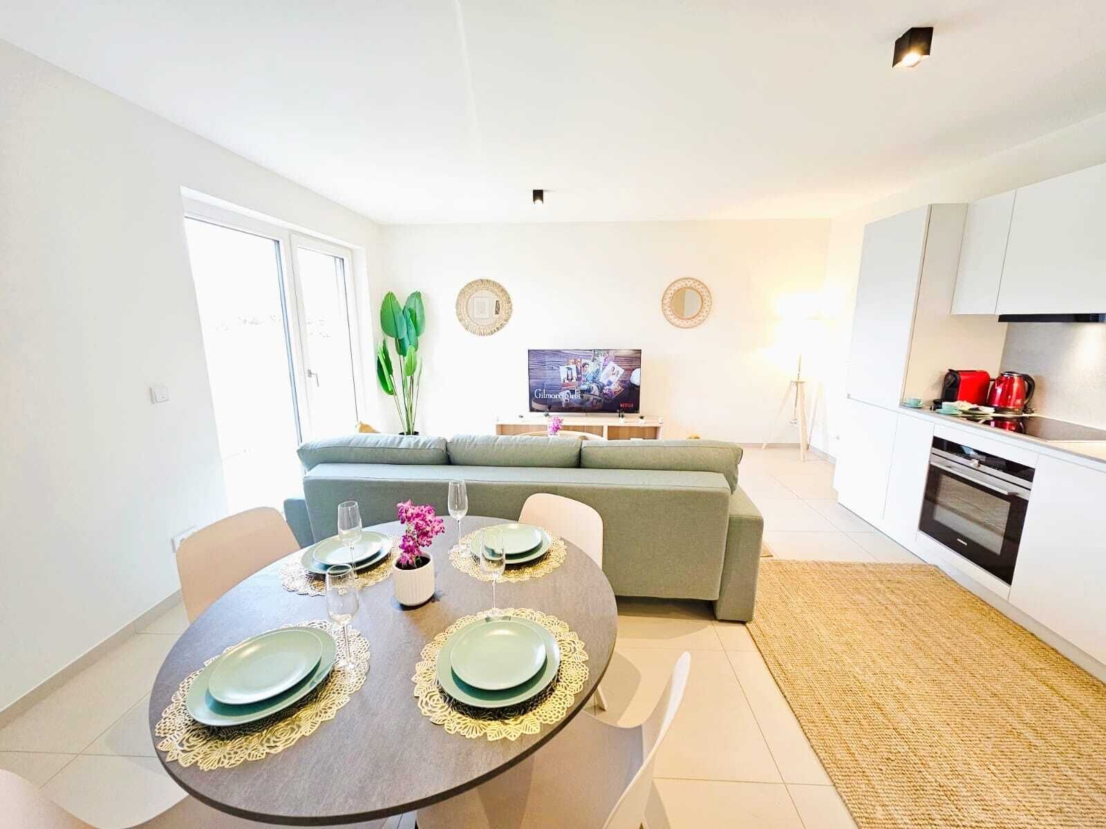 A modern living room with a dining table set for four, a grey sofa, a flat-screen TV, and a kitchen area with a stove and red appliances.