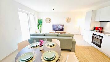 A modern living room with a dining table set for four, a grey sofa, a flat-screen TV, and a kitchen area with a stove and red appliances.