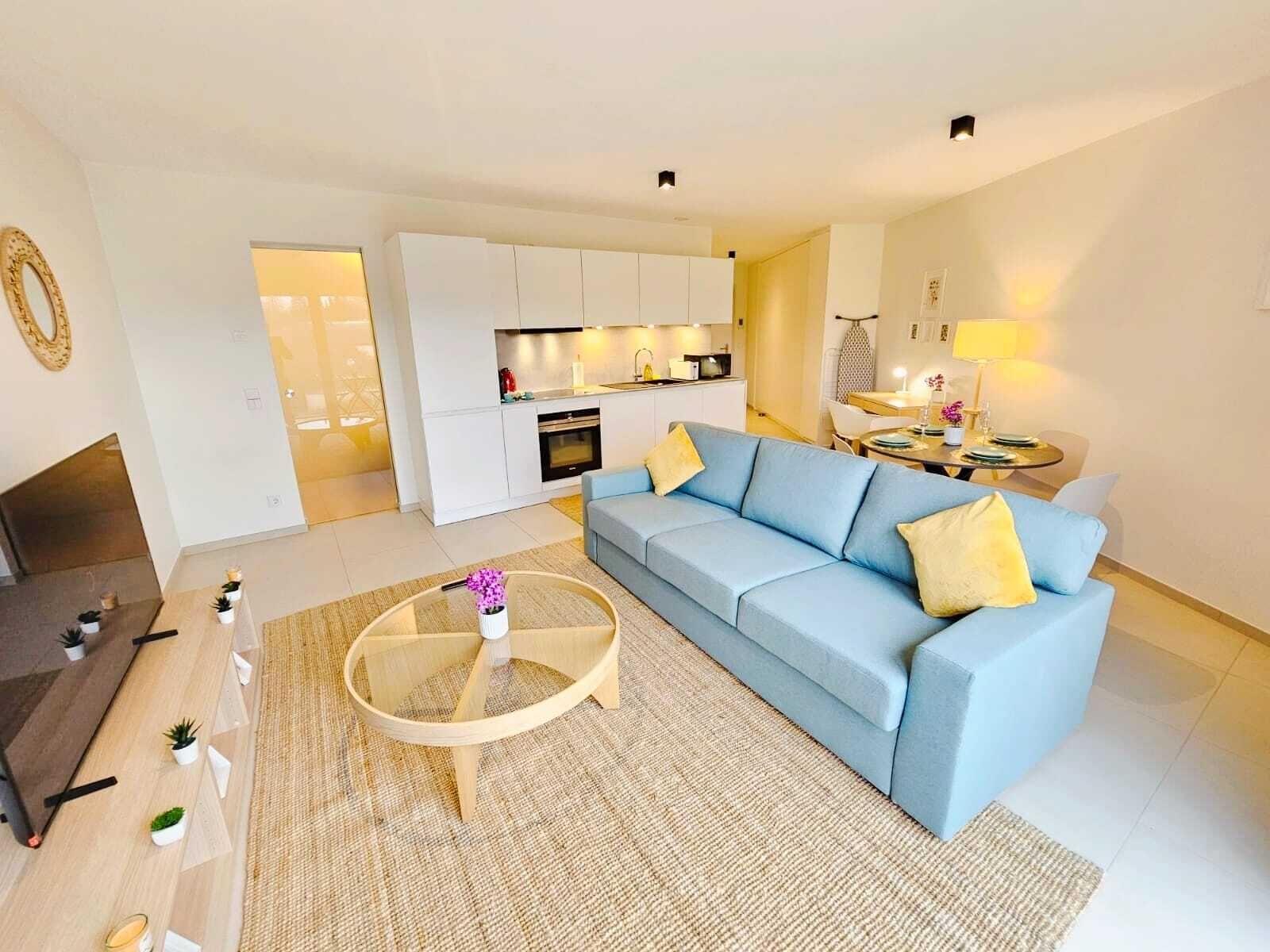 A modern living room with a light blue sofa, a glass coffee table, and a kitchen in the background.
