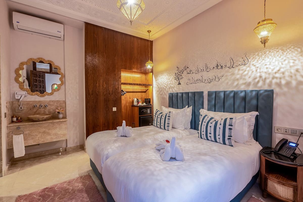 A hotel room with a large bed, a wooden wardrobe, a bathroom with a mirror and sink, and a wall with Arabic calligraphy.
