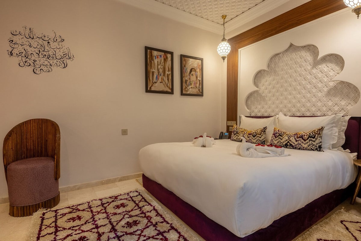 A bedroom with a large bed, a wooden chair, a decorative wall piece, and a patterned rug.