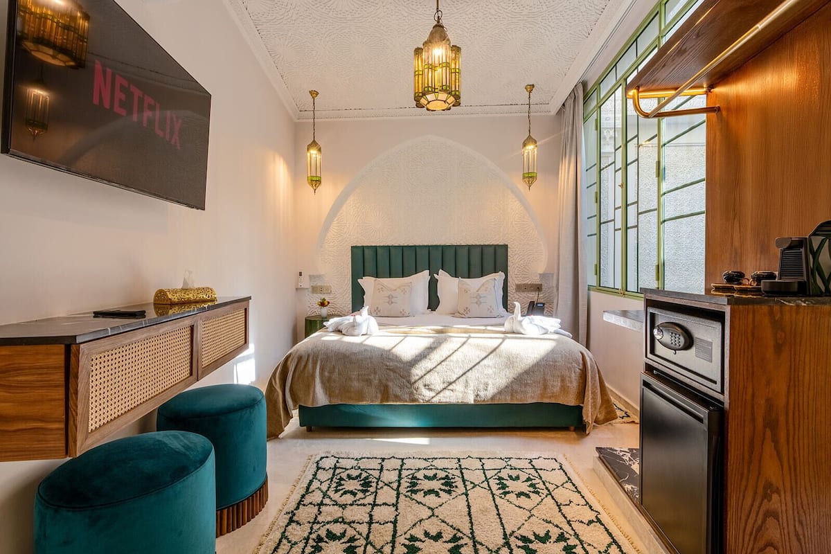 A bedroom with a bed, bedside tables, a desk, a TV, and a minibar.