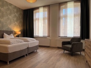 Ein Schlafzimmer mit einem Doppelbett, einem Sessel, einer Kommode und großen Fenstern mit Vorhängen.