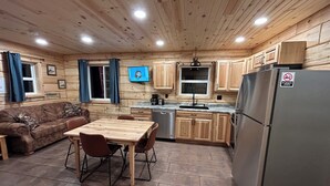 A cabin-style kitchen with wooden cabinets, a dining table with chairs, a refrigerator, and a television on the wall. - Cabin 1 on The Willow Reservoir (Hazelhurst)