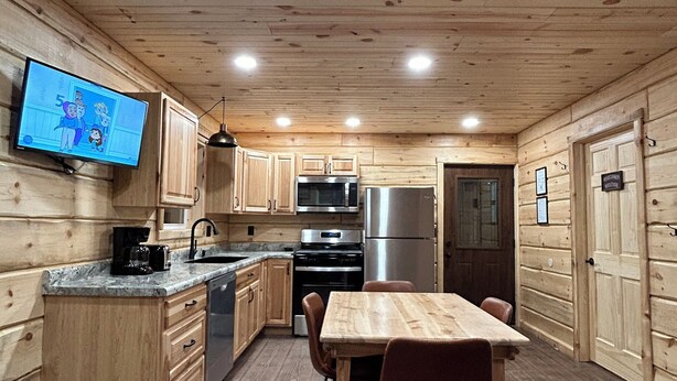 A wooden cabin interior with a kitchen area featuring a dining table, chairs, a TV mounted on the wall, and a refrigerator. - Cabin 1 on The Willow Reservoir (Hazelhurst)