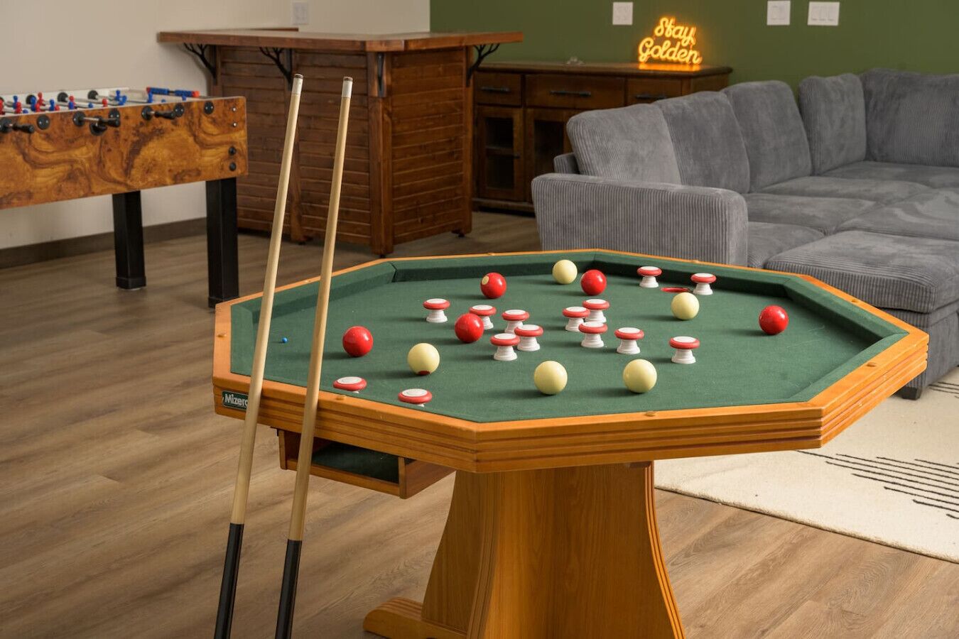Una mesa de billar con bolas, una mesa de futbolín y un sofá en una habitación con suelo de madera.