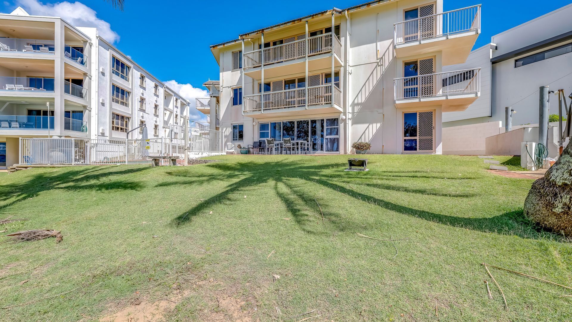 A multi-story residential building with balconies, a grassy courtyard, and a clear blue sky.