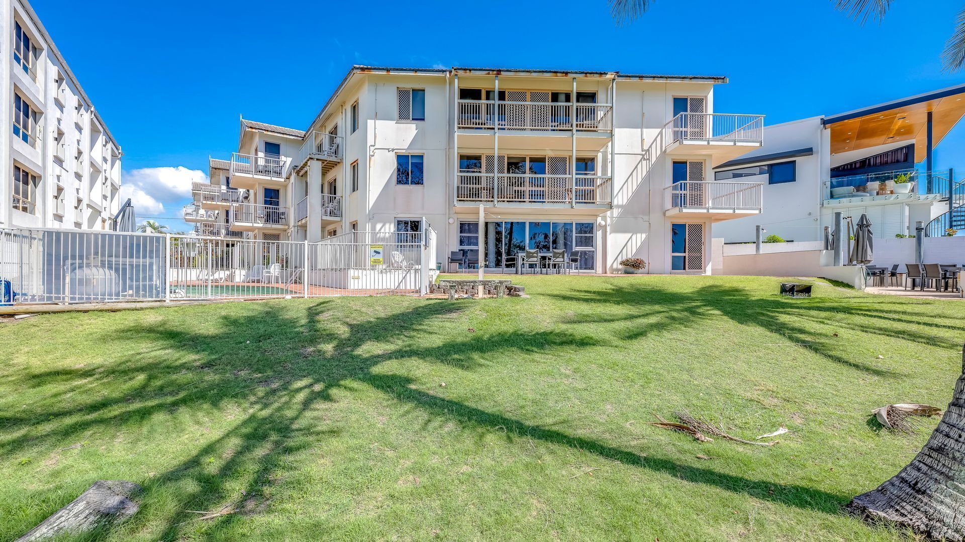 A modern multi-story residential building with balconies, a well-maintained lawn, and a clear blue sky.
