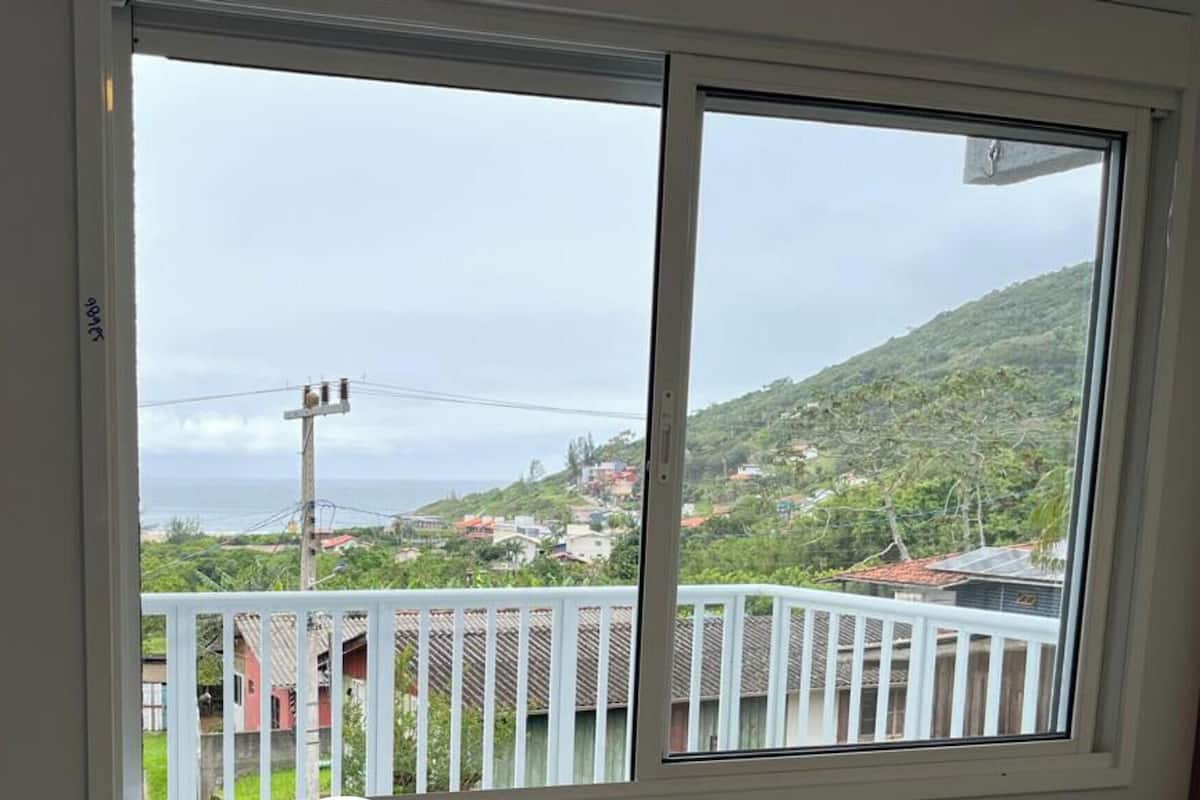 Een kamer met een groot raam dat uitkijkt op een balkon met uitzicht op een heuvel en de oceaan.