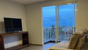 A living room with a TV on a wooden stand, a beige sofa, and a balcony with a view of the sea. - Paraíso na Gamboa - Garopaba - SC (Garopaba)