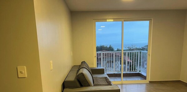A room with a sofa, a sliding glass door leading to a balcony with a white railing, and a view of the outside. - Paraíso na Gamboa - Garopaba - SC (Garopaba)