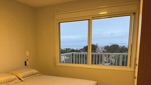 A room with a bed, a window with a view of a balcony and buildings, and a door. - Paraíso na Gamboa - Garopaba - SC (Garopaba)