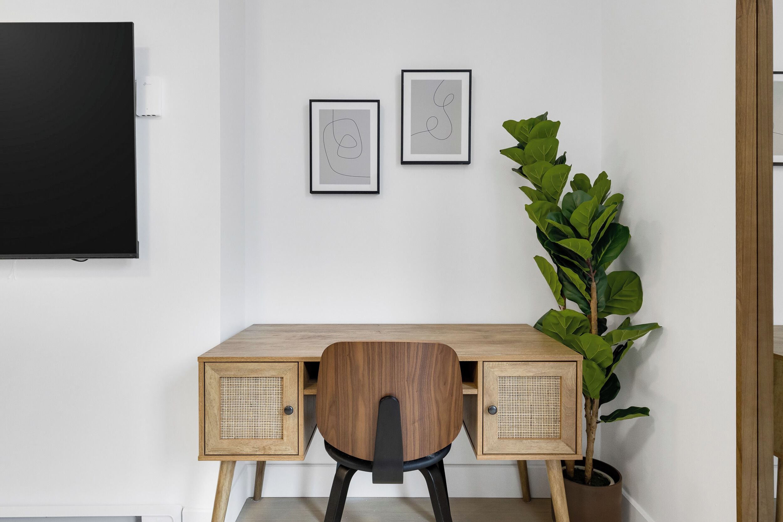 A modern workspace with a wooden desk, two framed abstract artworks, a large potted plant, and a black flat-screen TV mounted on the wall.