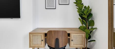 A modern workspace with a wooden desk, two framed abstract artworks, a large potted plant, and a black flat-screen TV mounted on the wall.