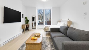 Un soggiorno moderno con un divano grigio, un tavolino in legno, una TV montata a parete, una scrivania con sedia e un'ampia finestra con vista sugli edifici.