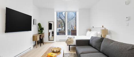 A modern living room with a grey sofa, a wooden coffee table, a TV mounted on the wall, a desk with a chair, and a large window with a view of buildings.