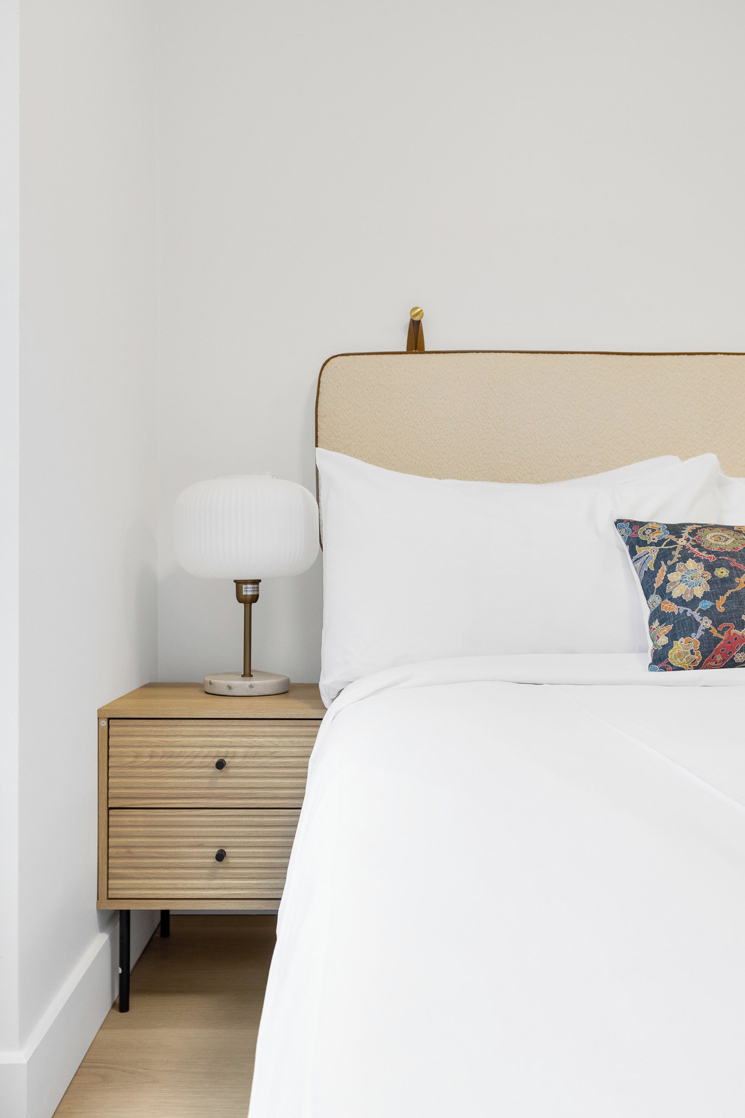 A neatly made bed with white bedding, a wooden nightstand with a lamp, and a colorful pillow.