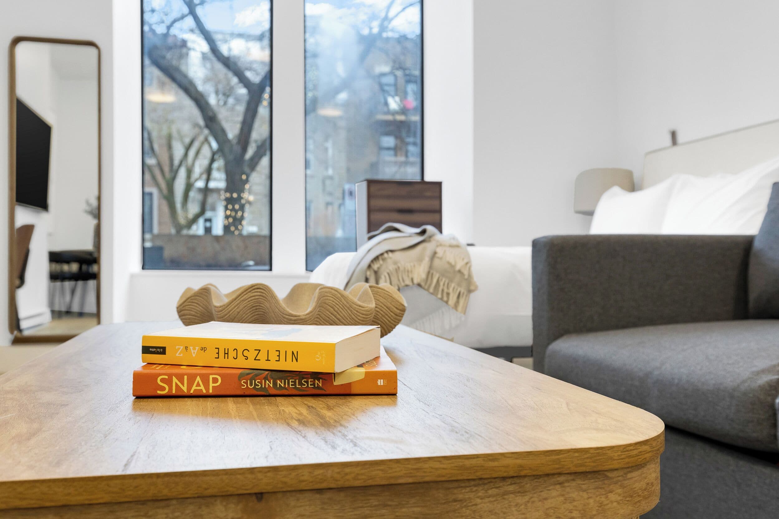 A wooden coffee table with books, a grey sofa, and a window with a view of buildings and trees.