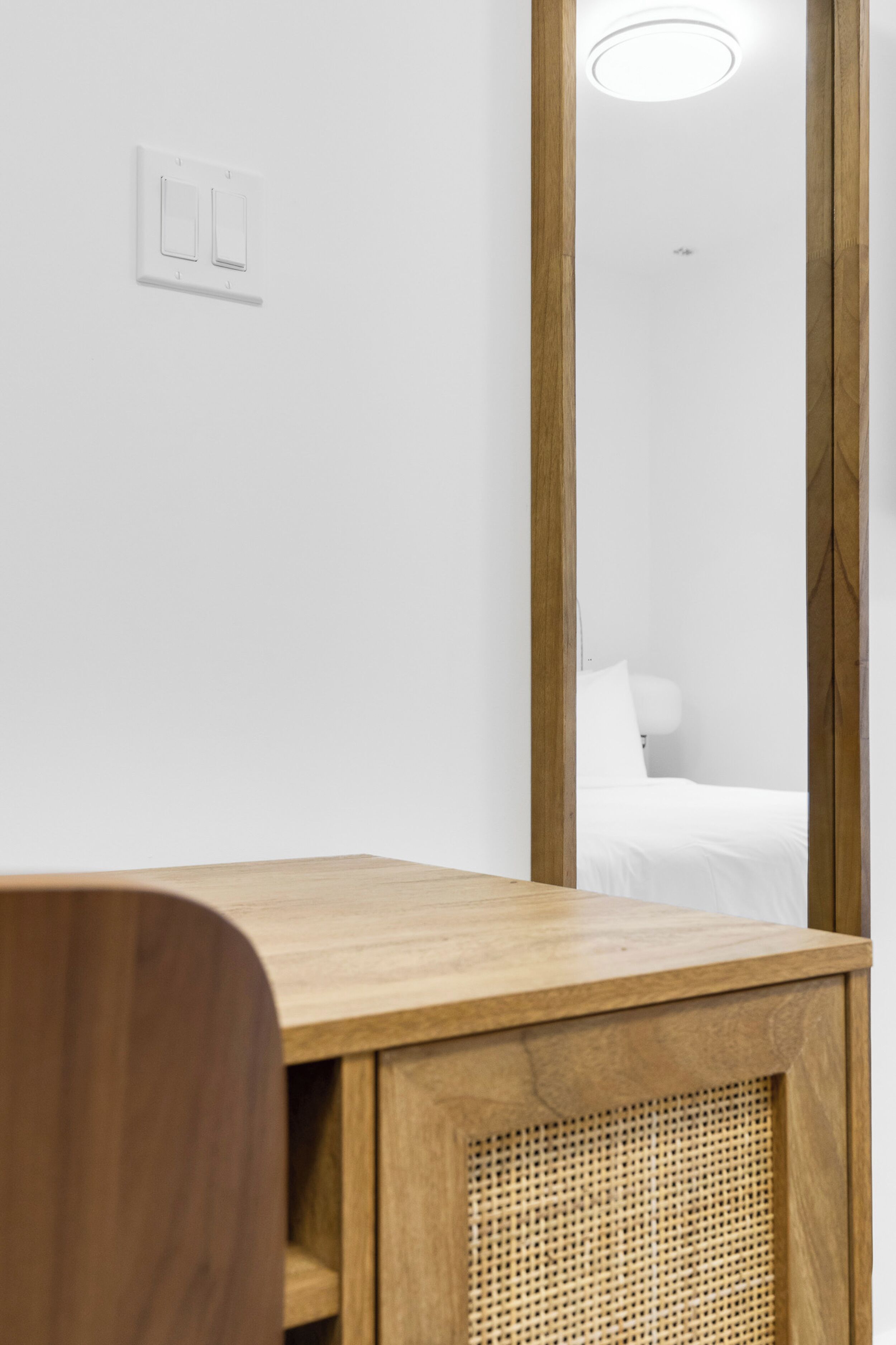 A wooden desk with a woven basket, a white wall with a light switch, and a mirror reflecting a bed with white bedding.