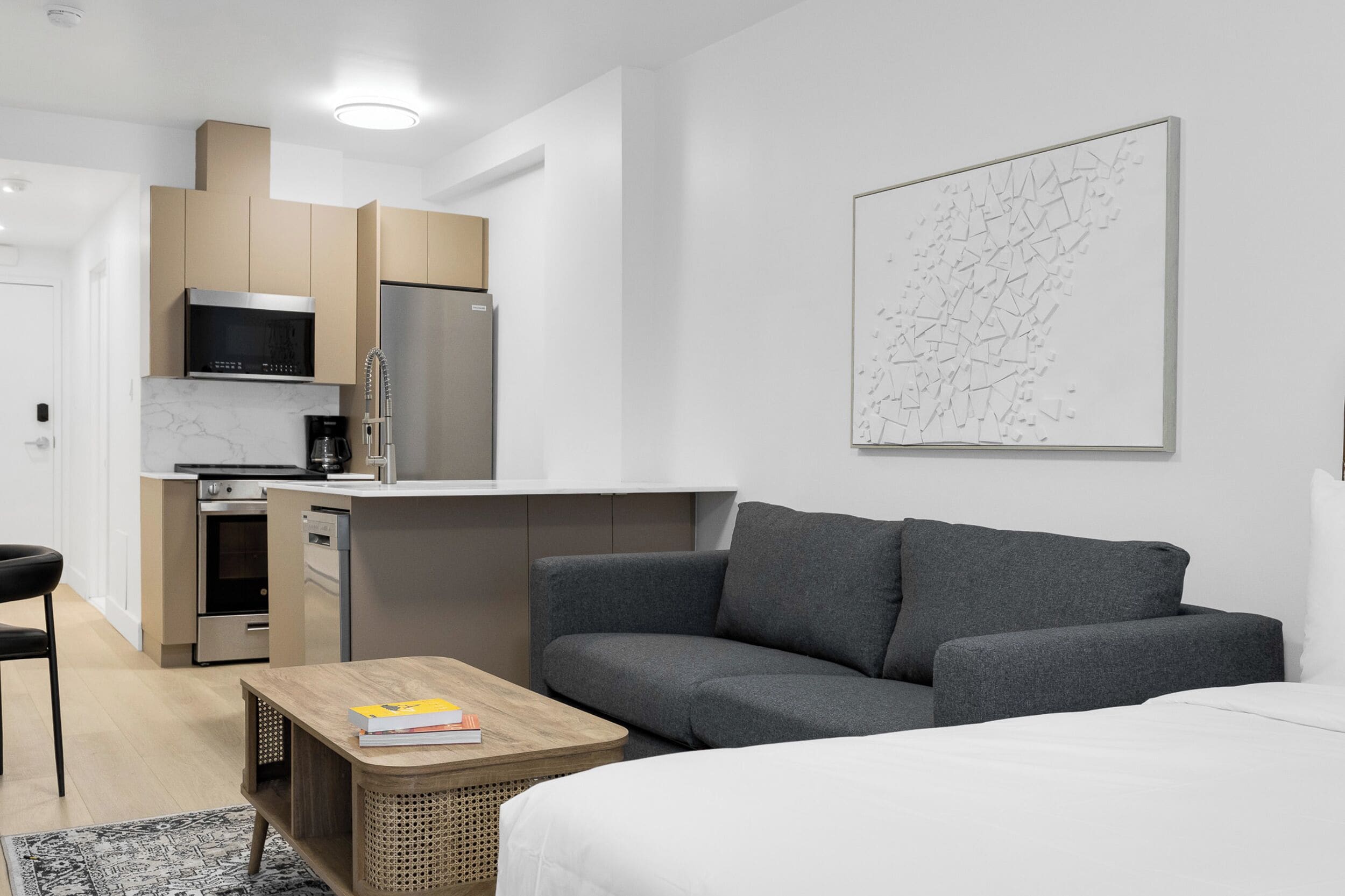 Ein modernes, kompaktes Wohnzimmer mit Küchenzeile, grauem Sofa und einem großen abstrakten Kunstwerk an der Wand.