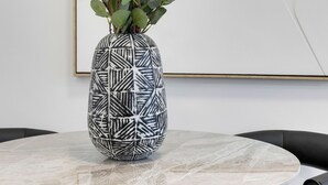 Un tavolo rotondo con un piano in marmo bianco, circondato da sedie nere. Sul tavolo è posata una composizione con un vaso a motivi bianco e nero e verde.