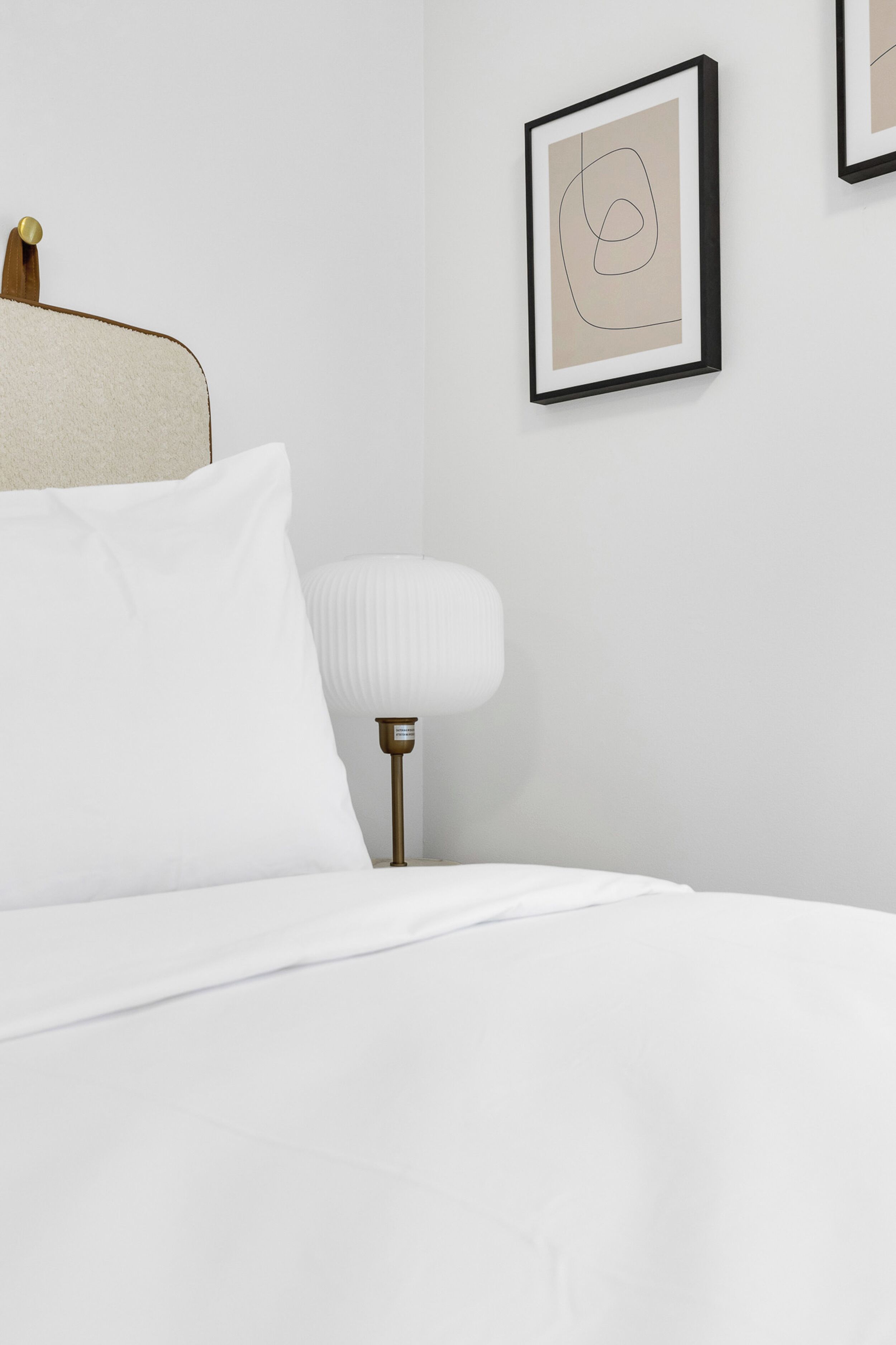 A neatly made bed with white linens, a white wall, and a framed abstract artwork.