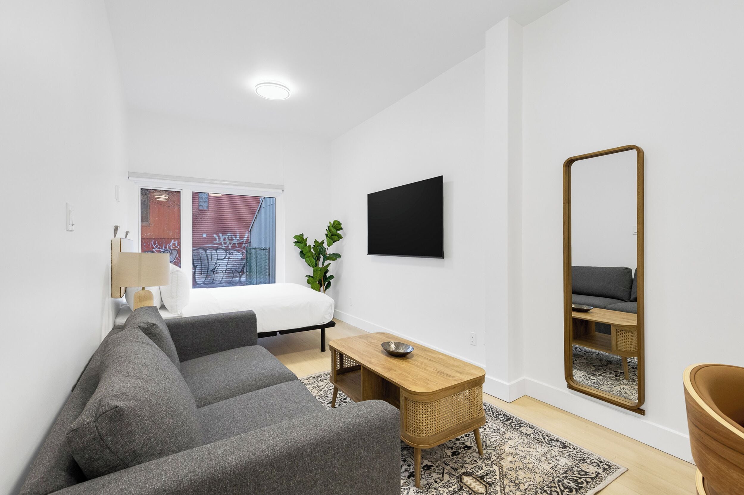 A modern, minimalistic living room with a grey sofa, a wooden side table, a large mirror, and a bed with a white headboard.