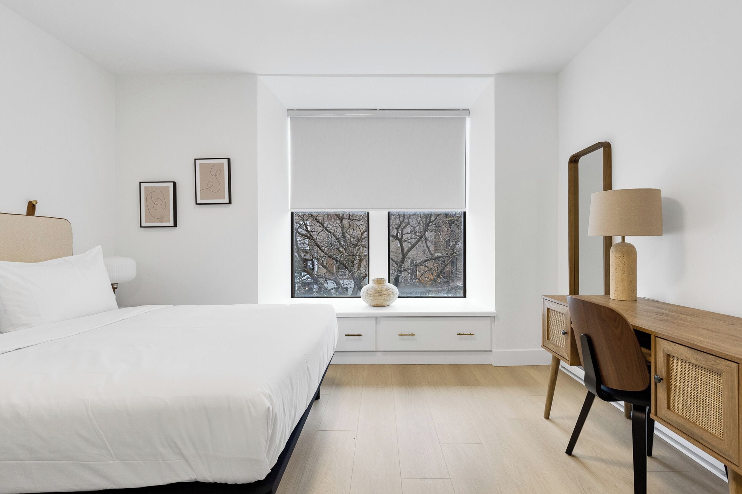 Um quarto com cama, escrivaninha com cadeira, luminária, janela com persianas e duas quadros na parede.