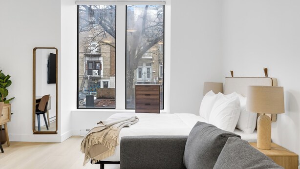 Una camera da letto moderna con una grande finestra, un letto con lenzuola bianche, un divano grigio, un comodino in legno con una lampada e uno specchio sulla parete.