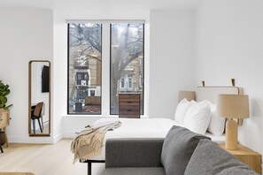 A modern bedroom with a large window, a bed with white linens, a grey sofa, a wooden nightstand with a lamp, and a mirror on the wall. - Le Mystral (Montréal)