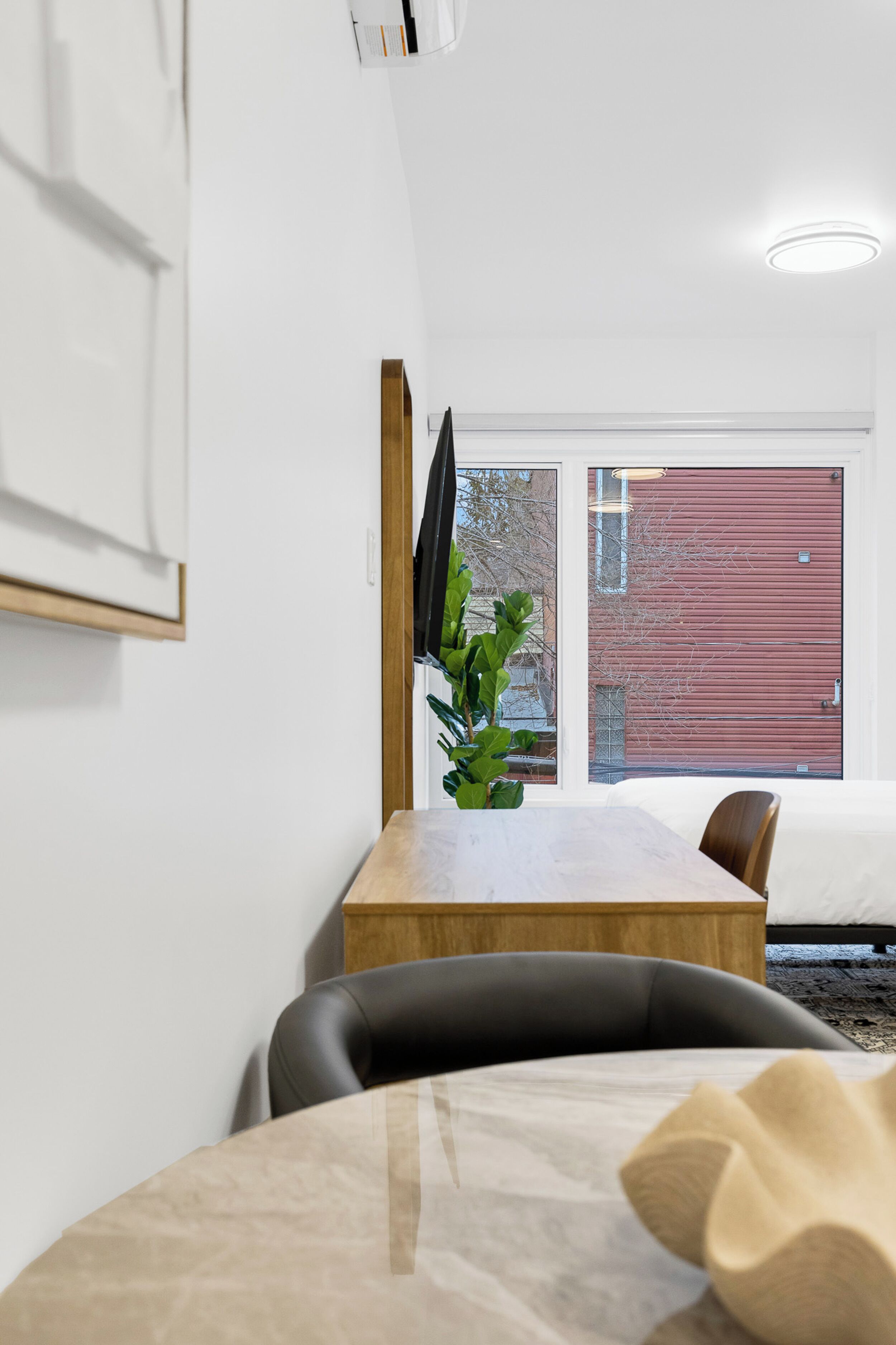 A modern dining area with a wooden table and chairs, a plant, and a view of a red building through a large window.