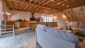 A spacious living area with a high ceiling, wooden beams, a kitchen with modern appliances, and a stone wall. - Le Grand Gite du Relais (PARGUES)