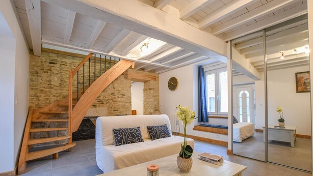 A spacious living room with a white sofa, a wooden staircase, and a stone wall. - Le Grand Gite du Relais (PARGUES)
