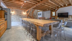 A spacious kitchen with a long wooden island, clear glass chairs, and a stone wall. - Le Grand Gite du Relais (PARGUES)