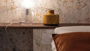 Une chambre avec un papier peint à motifs floraux, une lampe de chevet, un vase jaune et un coussin marron.