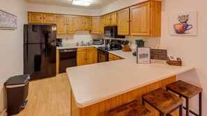 A modern kitchen with wooden cabinets, a stainless steel refrigerator, a microwave, and a coffee machine. There are two wooden stools at the counter. - Slope Views- Ski Shuttle- Pool & Hot Tub (Lincoln)