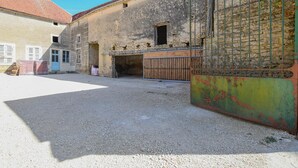 A courtyard with a building featuring a tiled roof, a metal gate, and a gravel ground. - Le Petit Gite du Relais (PARGUES)