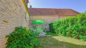 A stone building with a tiled roof, a green umbrella, and a small patio with chairs. - Le Petit Gite du Relais (PARGUES)