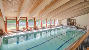 Una piscina interior con vigas de madera, agua cristalina y múltiples sillones de descanso a lo largo de la orilla.