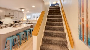 Una cocina moderna con una isla central, una escalera con barandillas de madera y vistas al salón.