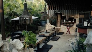 An outdoor seating area with tables and chairs, surrounded by tropical plants and a stone wall. - Siwa Jungle Solstice C1-102 (Tulum)